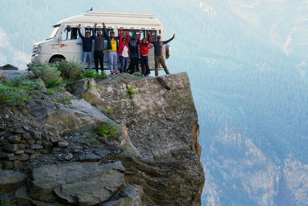 A group of friends celebrating on a cliff edge with a van in the background during a mountain adventure.